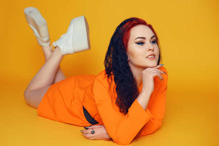 A woman with long curly hair lies on the floor against a yellow background. A girl in an orange dress with red hair and arrows in her eyes in white sneakers. Beauty and fashion. Summer sales.の写真素材