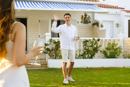 Couple celebrating buying a house by the ocean. couple drinks wine near the house. Couple rejoices, smiles, laughs. Beautiful girl in a white dress on a date with a man in white. Romantic travel.の写真素材