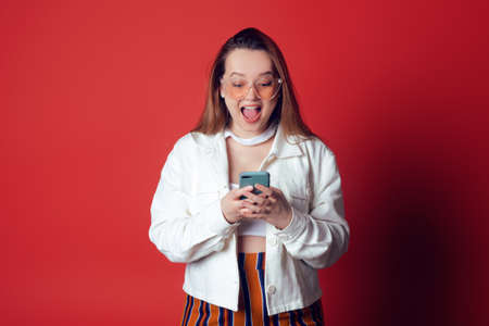 Beautiful, sweet, brunette on a red background in the studio. The girl looks at the phone in shock, joy, surprise, big wins or discounts. Empty space for text. Model in sunglasses, summer photo.の写真素材