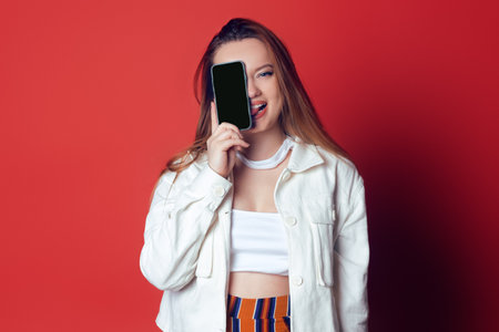 Beautiful, sweet, brunette on a red background. The girl holds the phone in front of her face and shows positive emotions. The model grimaces and shows the phone. Phone screen, empty space for text.の写真素材