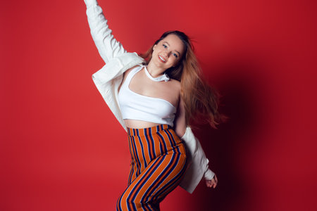 Beautiful energetic girl dancing in stylish things in the studio on a red background. Joy, happiness, dance, movement, fun. Woman smiles and looks at the camera, summer photo. Long hairの写真素材
