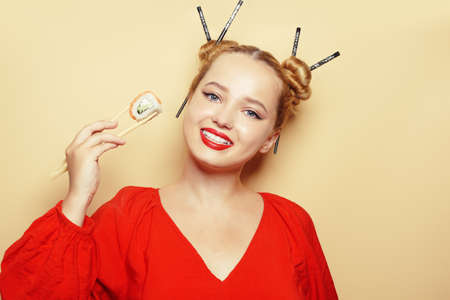 Beautiful girl eating sushi on a white plate. A woman in a red dress and red lips eats sticks of sushi with Philadelphia salmon. Model smiling and looking at the camera.の写真素材