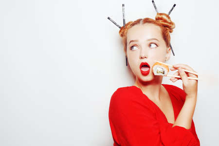 Beautiful girl eating sushi on a white plate. A woman in a red dress and red lips eats chopsticks of sushi with salmon and shrimp. Fashion photo models, sushi restaurantの写真素材