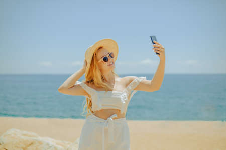 A beautiful girl on the background of the beach, sea and ocean makes a photo on the phone. Blonde in white outfit on vacation blogging, freelancer, vacation earnings, summer.の写真素材