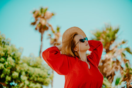 Fashion photo of a girl in black glasses with a chain and a large straw hat, a red dress on a background of palm trees. Wind in the hair of the model, summer photo, vacation, rest. The girl enjoys lifeの写真素材