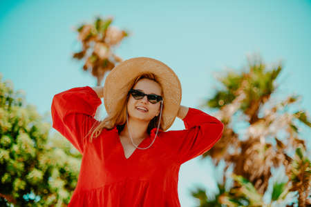 Happy girl in a red dress on vacation on the background of palm trees laughing and smiling. Vacation. A woman in sun glasses and a big hat enjoys life and dancesの写真素材
