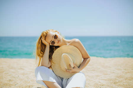 Fashion photo of a girl on the beach against the background of the sea or the sun, the girl covers her body with a hat. Beautiful woman with large earrings and in a white suitの写真素材