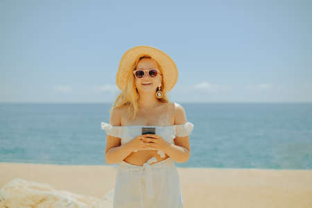 Beautiful girl on the background of the beach, sea and ocean. A blonde in a white outfit on vacation looks at the phone, roaming services, freelancer, income earning on vacation, summer internet applicationsの写真素材