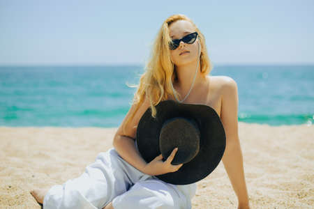 Fashion photo of a girl in black glasses with a chain and a large straw hat, against the backdrop of the sea or ocean. Woman sunbathes on the beach, summer photo. Blonde with long hairの写真素材