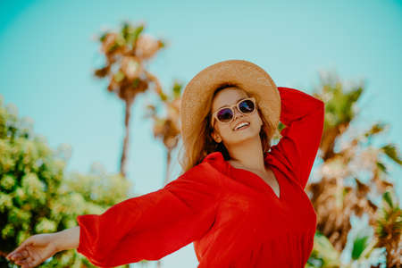 Happy girl in a red dress on vacation on the background of palm trees laughing and smiling. Vacation. Woman in sun glasses and big hat enjoys lifeの写真素材