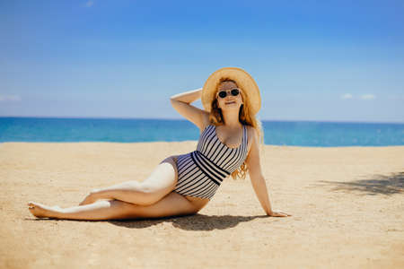 A beautiful blonde girl in a swimsuit and sun glasses and a large straw hat lies on the beach. Pretty woman sunbathes against the background of the sea, vacation, rest on the sand smileの写真素材