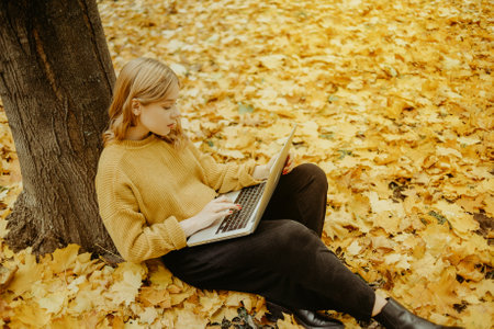 A beautiful girl teenager sits in an autumn park with a laptop. The student studies on the street. The girl looks into the laptop monitor and prints something.の写真素材