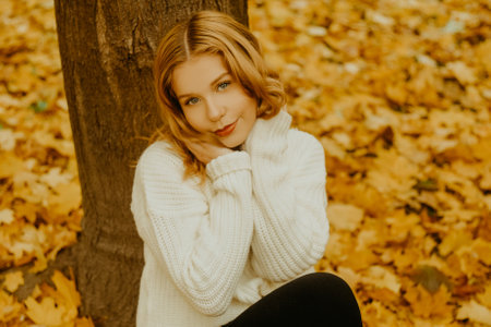 A girl in a white sweater sits on the autumn leaves. A beautiful blonde with a square looks at the camera. Woman outdoors in the autumn parkの写真素材