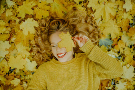 A beautiful young girl lies in the park with autumn leaves, a cute model with beautiful blond hair. Autumn discounts, autumn yellow sweater.の写真素材