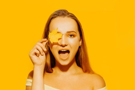 Girl with autumn leaves and red makeup, grimaces and has fun. Autumn photo on yellow backgroundの写真素材