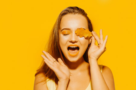 Girl with autumn leaves and red makeup, grimaces and has fun. Autumn photo on yellow backgroundの写真素材