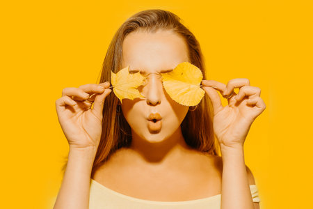 Girl with autumn leaves and red makeup, grimaces and has fun. Autumn photo on yellow backgroundの写真素材