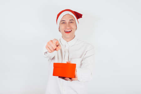 A guy in a Santa hat in the New Year is holding a gift in his hands, a red box. Man points his finger to the cameraの写真素材