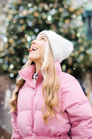 A cute portrait of a girl in the winter on the street, the snow is falling, the blonde on the background of the Christmas tree catches snowflakes with her tongue. Girl in a white hat, pink jacket, new year, smileの写真素材