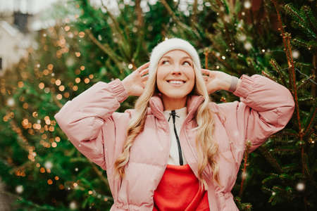 A cute blonde in a hat and a pink jacket stands against the background of Christmas trees. A girl buys a Christmas tree, New Year's mood, winter, holidays, New Year smileの写真素材