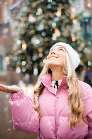 Cute portrait of a girl in the winter on the street, the snow is falling, the blonde on the background of the Christmas tree catches snowflakes. Girl in a white hat, pink jacket, new year, smileの写真素材