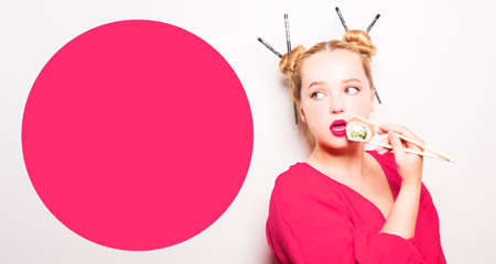 Woman in pink kimono eats philadelphia sushi with chopsticks, enjoys. Red lipstick and Japanese hairstyle, space for text. Pretty girl looks at the text, fashion.の写真素材
