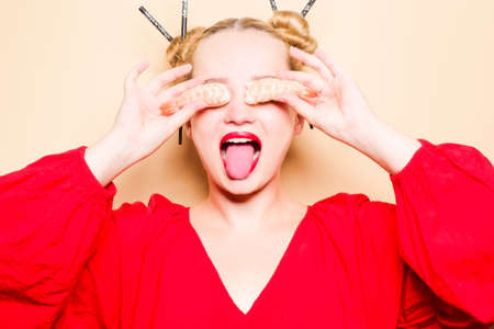 Girl in a red dress on a beige background. A woman eats with chopsticks for sushi, closes her eyes with them and grimaces, smiles.の写真素材