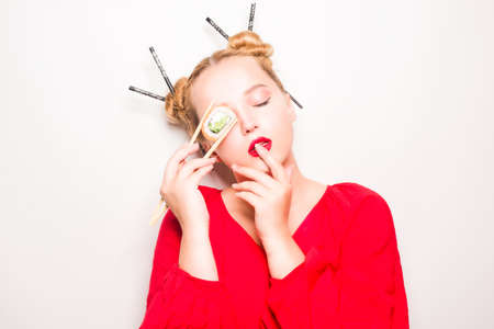 Woman in red kimono eats philadelphia sushi with chopsticks, enjoys. Red lipstick and Japanese hairstyle, space for text. Beautiful girl fashion photo on a white background.の写真素材
