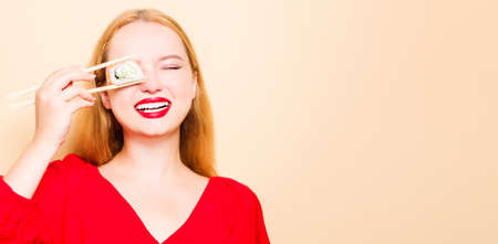 Girl in a red dress on a beige background. A woman eats with chopsticks for sushi, closes her eyes with them and grimaces, smiles place for textの写真素材