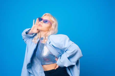 A girl in a blue jacket and purple glasses poses on a blue background and dances. Fashion photo in style 80s 90s party. Blonde girl in a shiny sweater and long earrings.の写真素材
