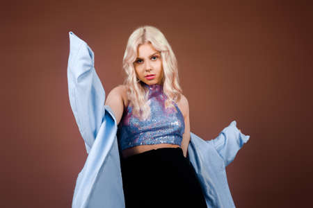 A blonde in a turquoise jacket, in a blue jacket in sequins, poses in the studio on a brown background. High fashion, 90s 80s. The girl dances and moves. Model with pink makeup.の写真素材