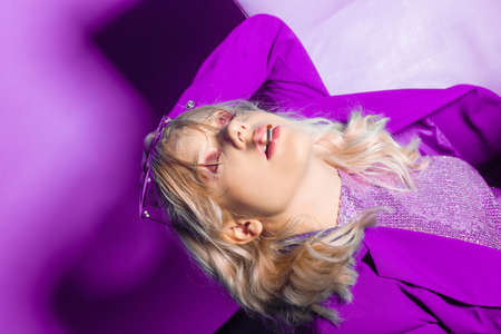 Incredibly fashionable blonde woman in a purple jacket and glasses in the style of 80s 90s sits on an armchair and poses. On a purple background. Fashionable woman smokingの写真素材