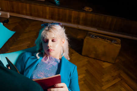 The girl is like in the clip from the 80s 90s. Short blonde haircut. The girl is sitting on a chair in a blue jacket and a shiny jacket, glasses. Fashion & Style book in handの写真素材