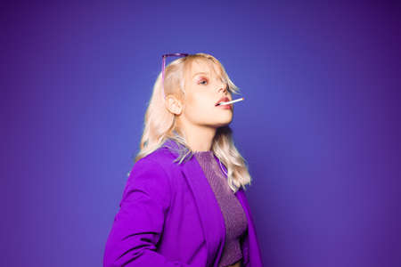 Incredibly fashionable blonde woman in a purple jacket and glasses in the style of 80s 90s poses. On a purple background. Fashionable woman smokingの写真素材