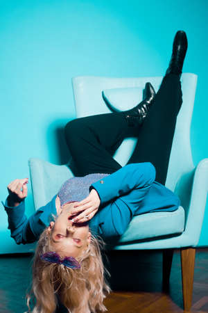 Incredibly fashionable blonde woman in a blue jacket and glasses in the style of 80s 90s sits on an armchair and poses. On a blue background. Fashionable woman smoking.の写真素材