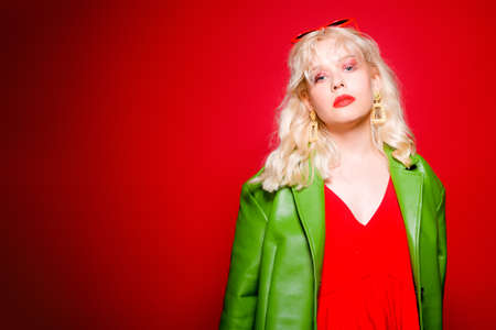 Blonde, high fashion, woman in earrings, bijouterie. Bright pink glitter makeup. Red glasses from the 90s and a stylish green raincoat, dress. On a red background.の写真素材