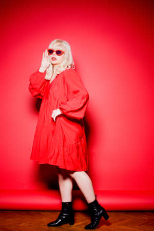 The blonde jumps on a red background in stylish red glasses and a dress. Girl model, dancing and posing, bright photo of fashion and style, black bootsの写真素材