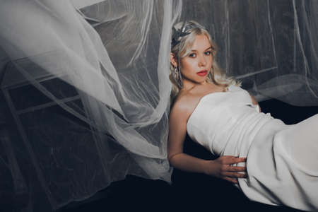 A girl in a wedding dress and a silver crown is lying on a sofa. Blonde queen. Fashion photo of a model looking at the camera.の写真素材
