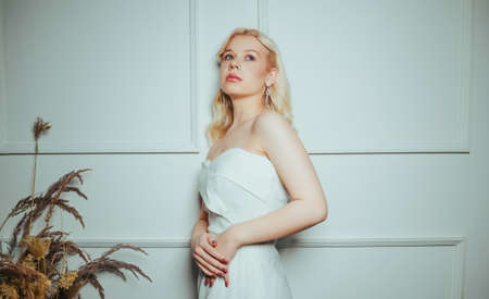 The bride is standing against the wall, a chic dress. Salon of wedding dresses. Woman in white, long earrings. Beautiful fashion blonde.の写真素材