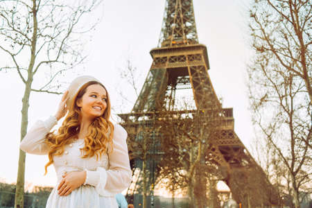 A girl against the backdrop of the Eiffel Tower in Paris in a beret and a dress with curled hair, a romantic journey. Woman laughing and looking away.の写真素材