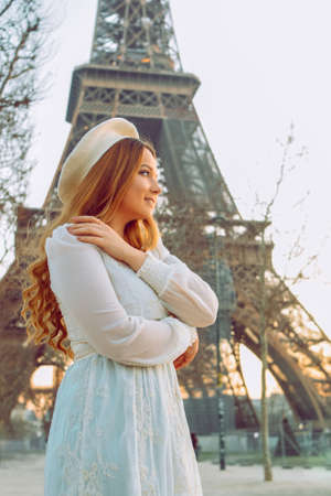 A girl against the backdrop of the Eiffel Tower in Paris in a beret and a dress with curled hair, a romantic journey. Woman laughing and looking away.の写真素材
