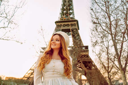A girl against the backdrop of the Eiffel Tower in Paris in a beret and a dress with curled hair, a romantic journey. Woman laughing and looking away.の写真素材