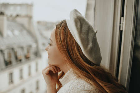 A woman stands near the window against the backdrop of the roofs of Paris. A girl with long hair in a beige suit and beret enjoys the city looking out the windowの写真素材