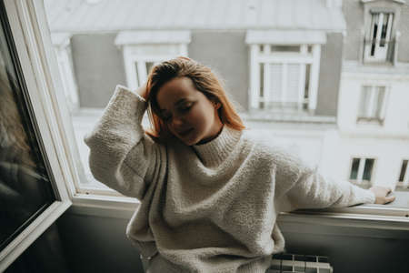 A woman stands near the window against the backdrop of the roofs of Paris. Girl with long hair in a beige suit posing.の写真素材