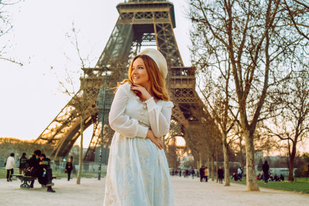 A girl against the backdrop of the Eiffel Tower in Paris in a beret and a dress with curled hair, a romantic journey. Woman laughing and looking away.の写真素材