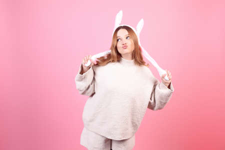 A beautiful girl in rabbit ears on a pink background grimaces, Easter holidays. The woman grimaces, kiss. Looks to the side, space for text.の写真素材
