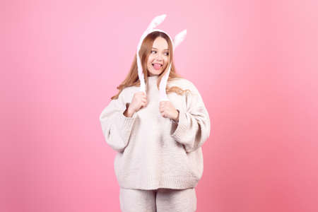 Beautiful girl in rabbit ears on a pink background laugh, Easter holidays. The woman smiles. Looks to the side, space for text, shows tongueの写真素材