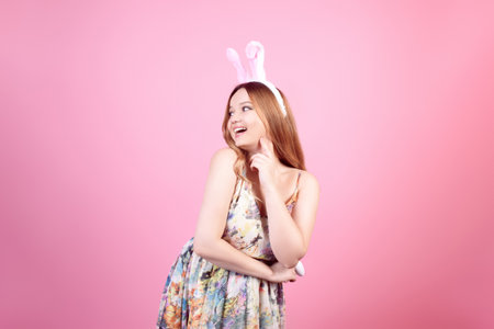 Girl in a dress with rabbit ears on a pink background, Easter holidays. Woman beautiful with long hair, easter eggs, smile and joy.の写真素材