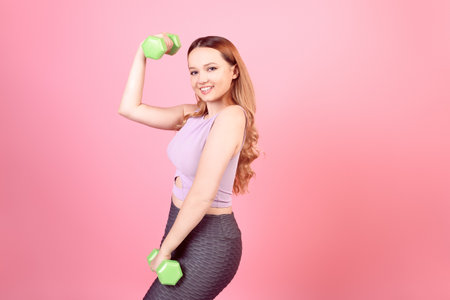 A beautiful girl goes in for sports on a pink background. The girl holds weights and shows exercises on her hands, sports goods, a healthy lifestyle. Woman in leggings.の写真素材