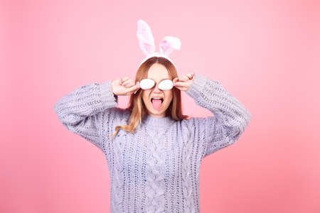 Beautiful girl in a blue sweater on a pink background. Woman with bunny ears, posing for easter, covering her eyes with easter eggsの写真素材